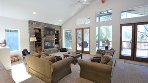 A spacious living room with a stone fireplace, large windows, and a ceiling fan. - Lake Shore - Lake Front (Bass Lake)