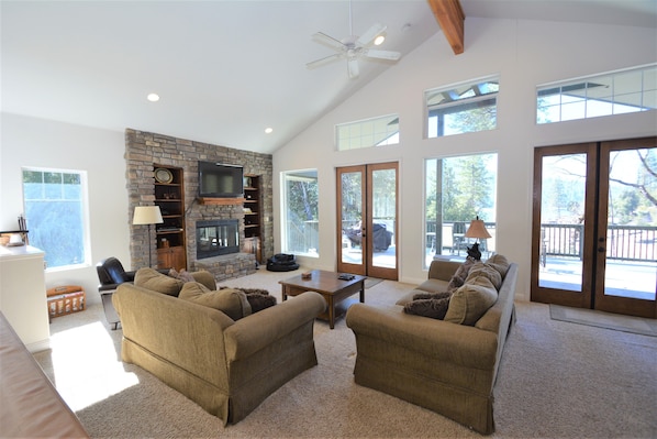 A spacious living room with a stone fireplace, large windows, and a ceiling fan.