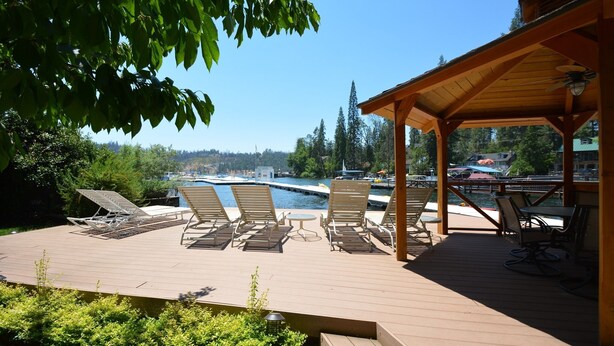A wooden deck with lounge chairs and a table, overlooking a pool and a scenic view of trees and buildings. - Johnson Home Lake Front, Boat slip, walking distance to Pines. Takes Pets (Bass Lake)