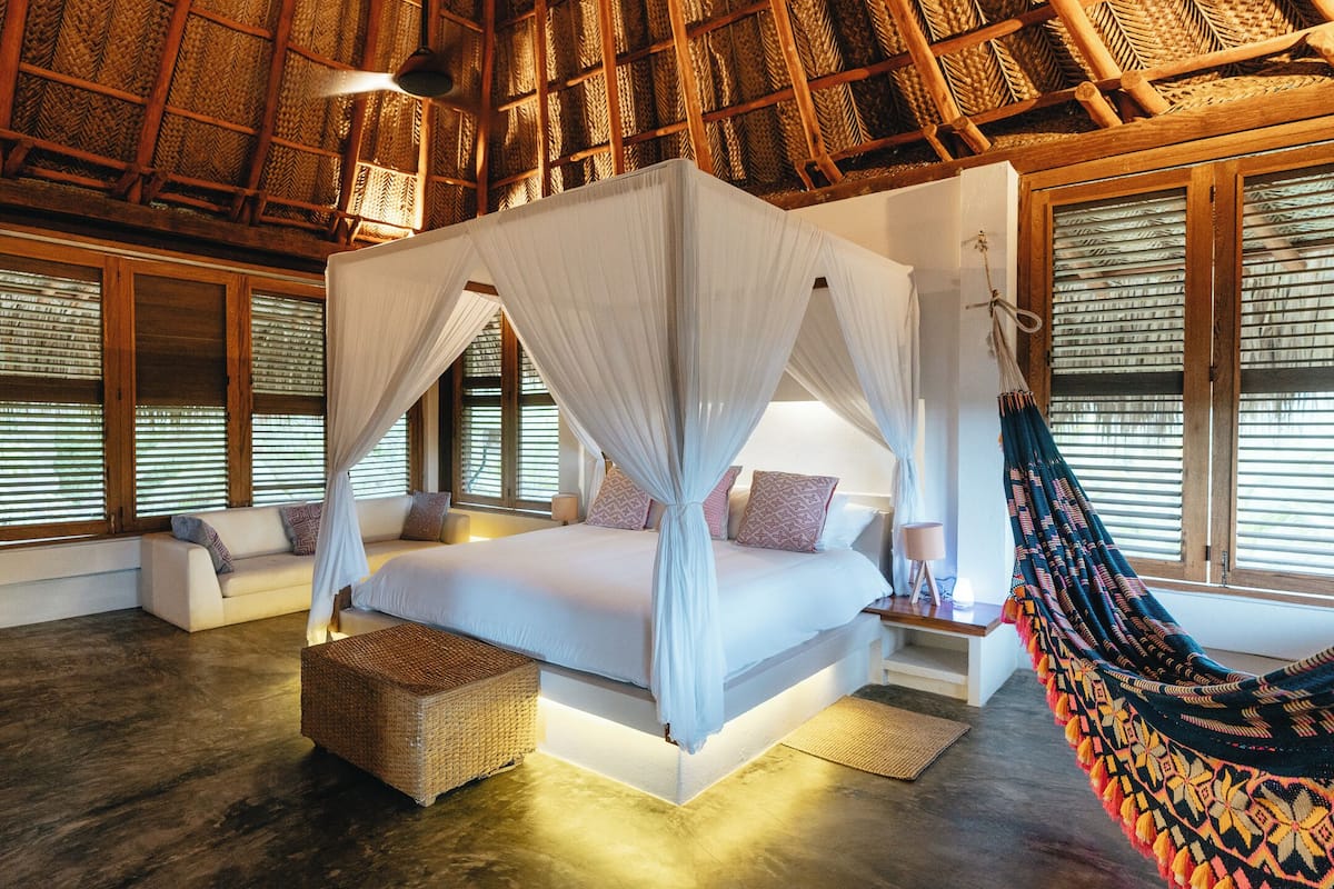A spacious bedroom with a large bed, a hanging hammock, and a view of the outside through large windows.