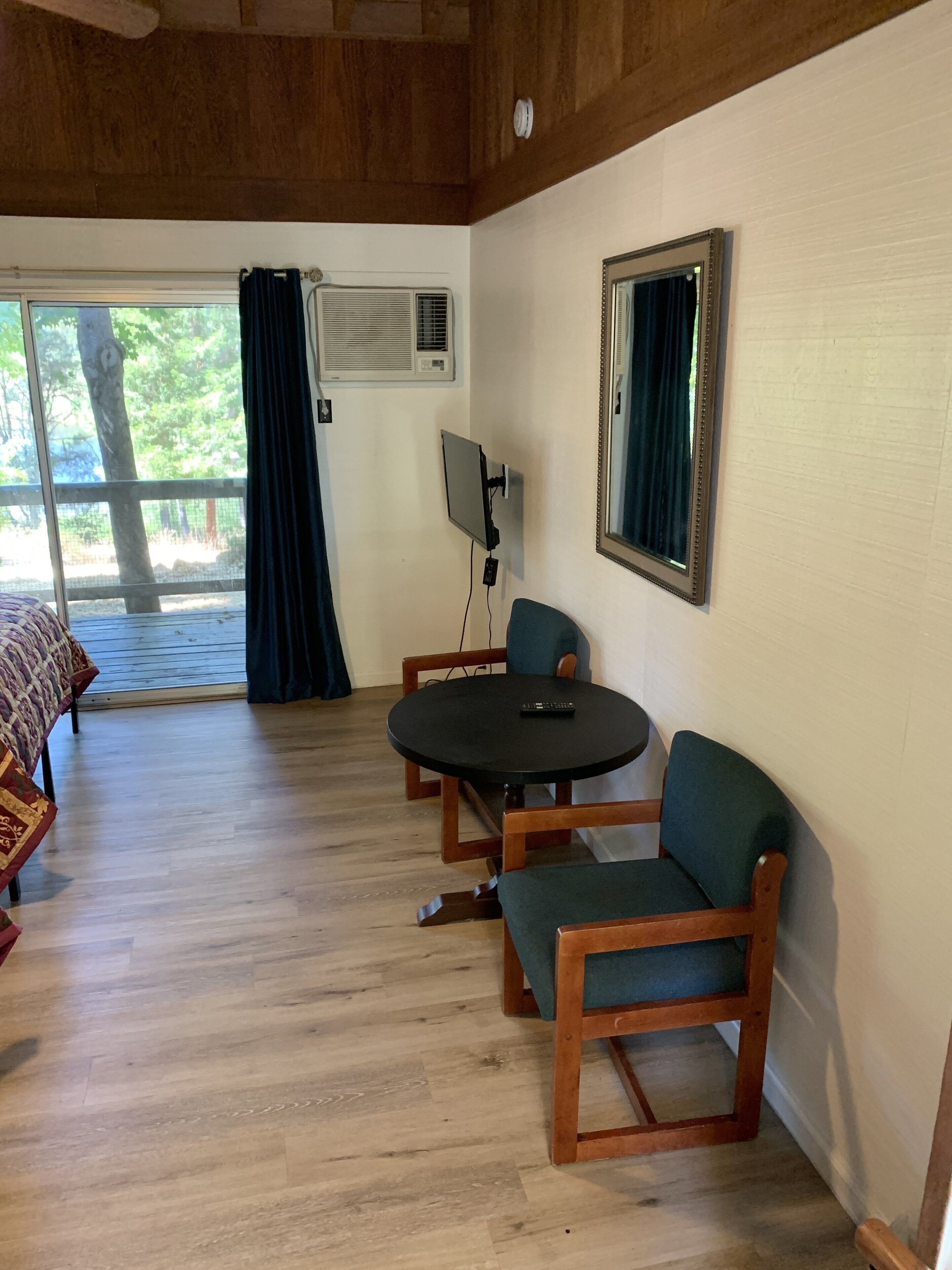 Ein Zimmer mit Holzboden, einem runden Tisch mit Stühlen, einem an der Wand montierten Fernseher und einem großen Fenster mit Vorhängen.