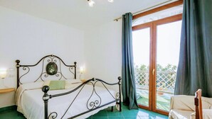 A bedroom with a metal bed frame, a bedside table with a lamp, a window with curtains, and a view of a balcony.