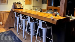 A kitchen with a wooden countertop, four white bar stools, a refrigerator, and a chalkboard with writing.