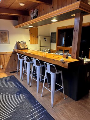A kitchen with a wooden countertop, four white bar stools, a refrigerator, and a chalkboard with writing.