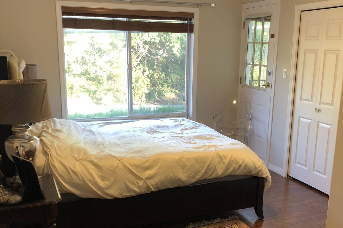 A bedroom with a large bed, a nightstand, a desk, a chair, a window with blinds, a door, and a patterned rug.