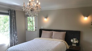 A bedroom with a large bed, a chandelier, a nightstand with a vase of flowers, and a door with a view of greenery outside. - Royalty B&B (Richmond)