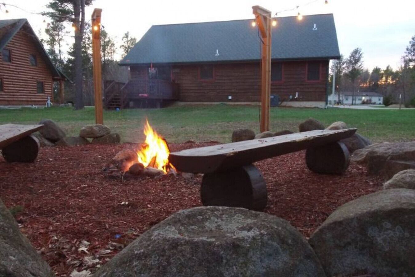 A wooden bench with a fire in the center, surrounded by rocks, in front of a cabin with a sloped roof.
