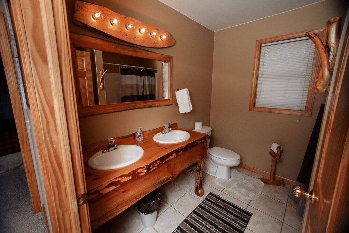 A bathroom with two sinks, a toilet, a mirror with lights, and a window with blinds.