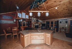 A wooden interior with a central island counter, wooden chairs, a stone fireplace, and a sign for 'KIMS Lakeside Cabins'.