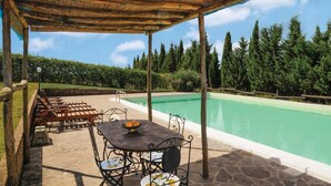 Ein überdachter Außenbereich mit Pool, Steinböden, Metallstühlen, einem Tisch mit Obstschale und Blick auf Bäume und klaren Himmel.
