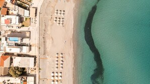 Vue aérienne d’un hôtel situé en bord de mer, avec des rangées de transats et de parasols, une piscine et des bâtiments adjacents.