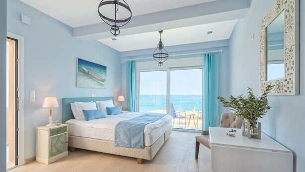 Une chambre à coucher comprenant un lit, des tables de chevet, un bureau, une chaise, une grande fenêtre donnant sur la mer et un luminaire au plafond.