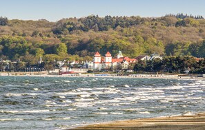 A coastal town with a prominent castle-like building with red roofs and a tower, situated by the sea with waves crashing onto the shore. - Prora Solitaire Panorama 01 (Binz)