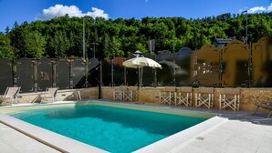 Piscina con sillas de descanso y sombrillas, rodeada de un muro de piedra y árboles.