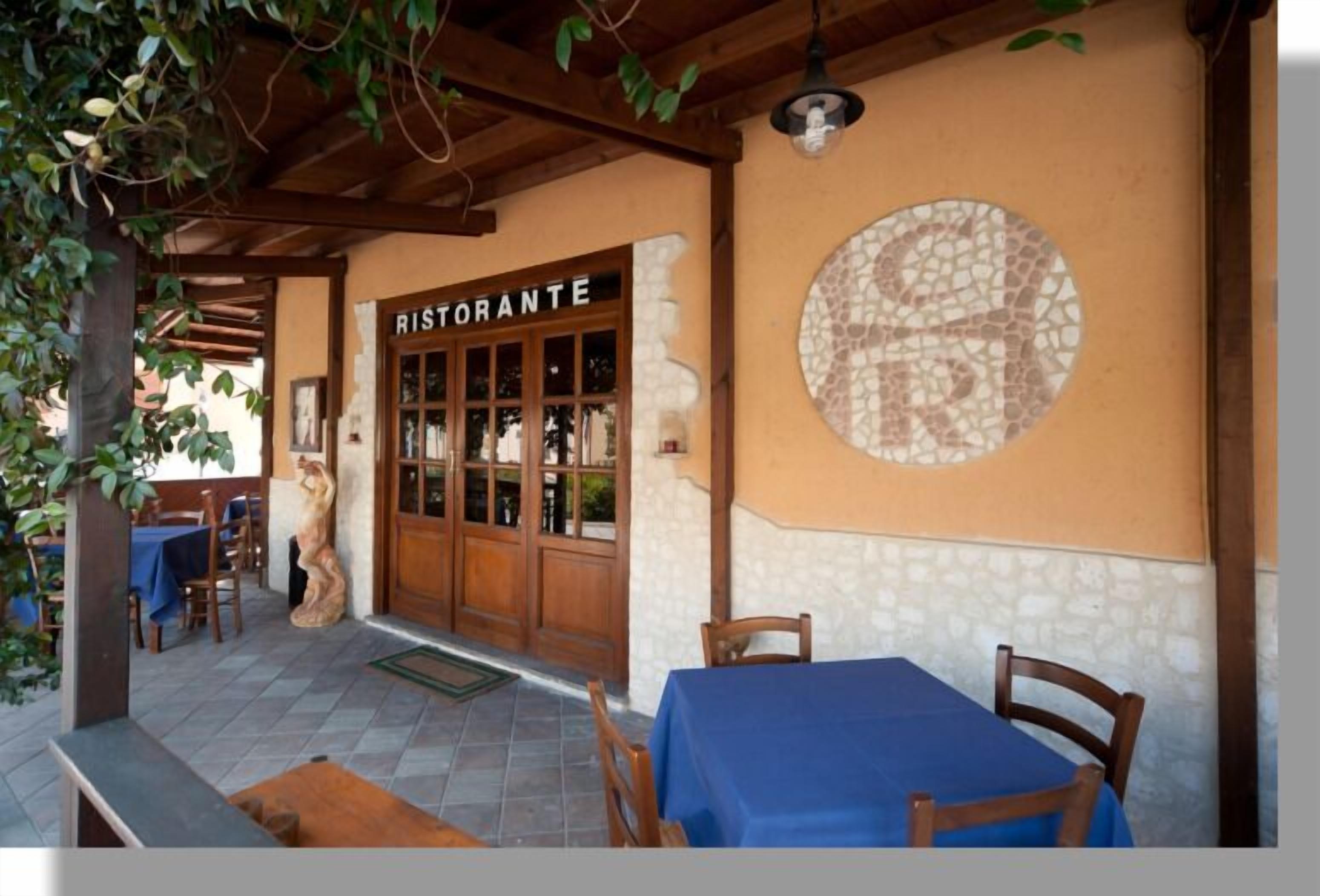 Un espacio de comedor al aire libre con mesas y sillas, una pérgola de madera y un muro de piedra con mosaico.