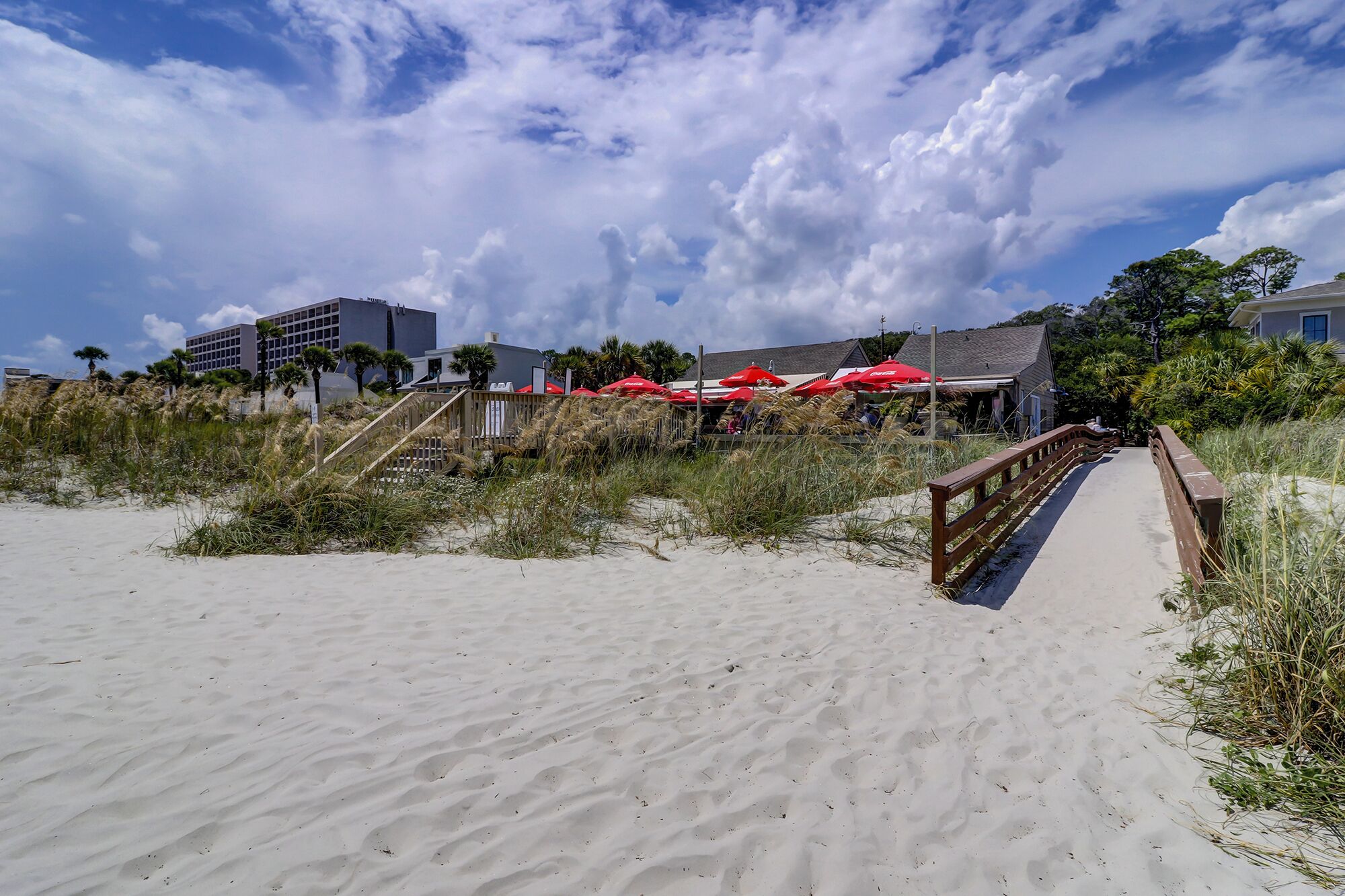 A beach with a wooden boardwalk leading to a building with red umbrellas.