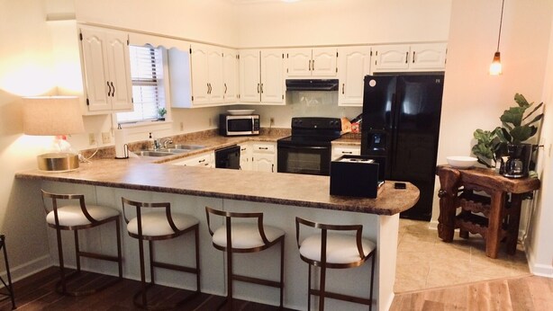 Una cocina con una isla central, taburetes de bar, armarios blancos, un refrigerador negro y piso de madera.