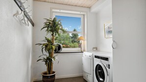 A laundry room with a washing machine, a potted plant, a window with a view of trees, and a wall-mounted coat rack.