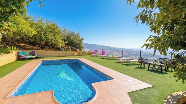 Une piscine rectangulaire dont le bord est recouvert de carreaux, entourée de gazon synthétique et agrémentée d’un patio avec des meubles d’extérieur.