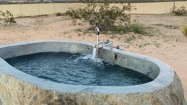 A stone hot tub with a faucet, surrounded by desert landscape and mountains in the background.