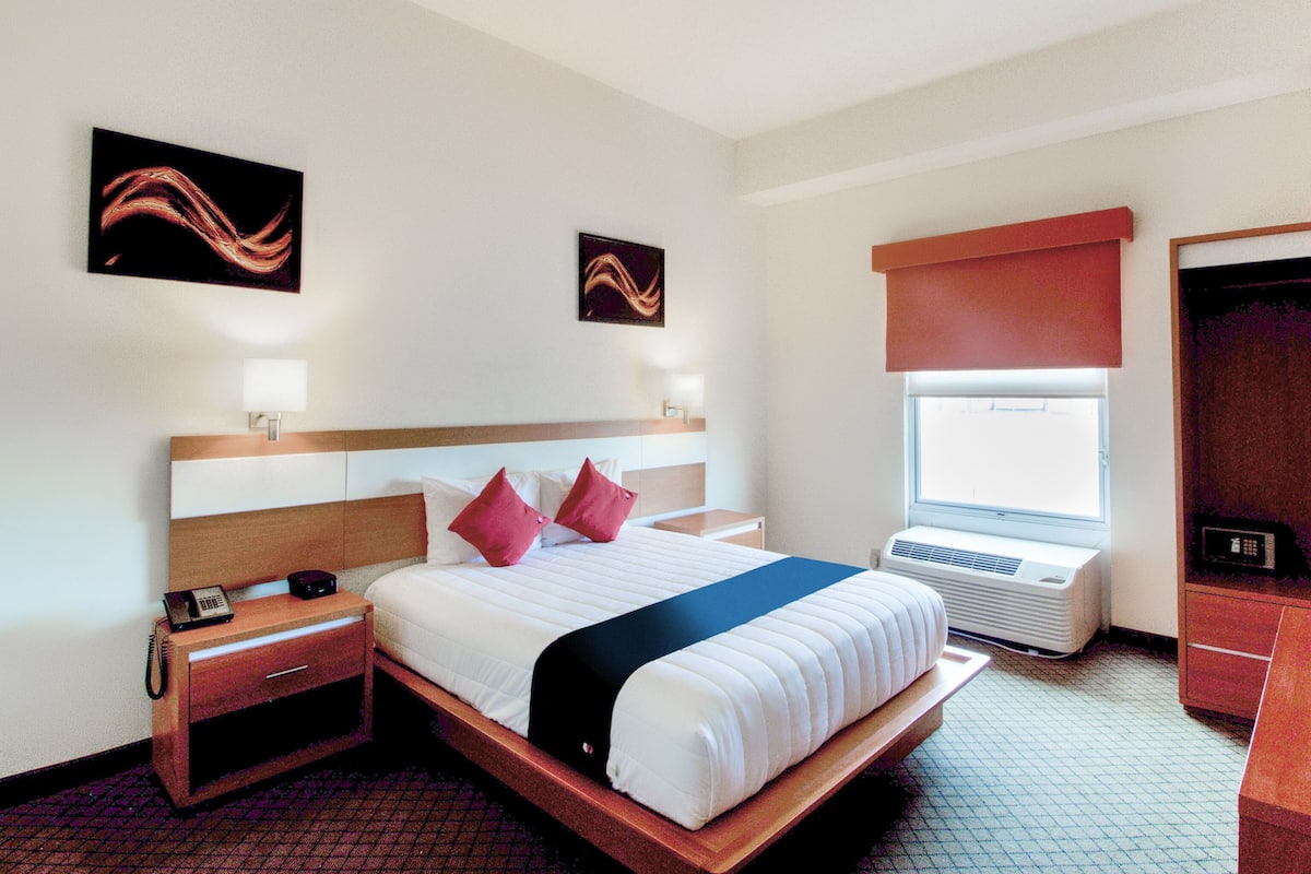 A hotel room with a bed, bedside table, lamp, artwork, window, air conditioning unit, and a wardrobe.