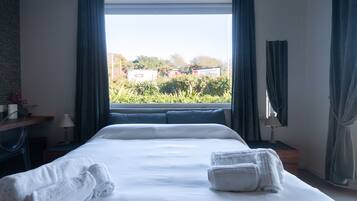 A hotel room with a large bed, two towels, a desk with a lamp, and a view of greenery and buildings outside.