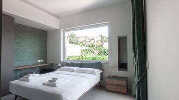 A modern bedroom with a large bed, a window with a view of greenery, a mirror, and a bedside table.
