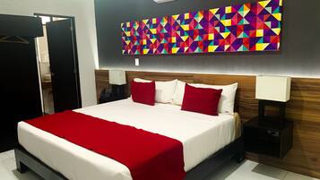 A hotel room with a large bed, red and white bedding, a colorful geometric wall art, a wooden headboard, a bedside table with a lamp, and a black wardrobe.