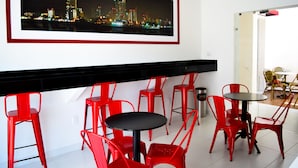 A modern dining area with red chairs and black tables, a framed cityscape picture on the wall, and a glimpse of a dining area with yellow chairs in the background. - Hotel Dwana (Mazatlán)