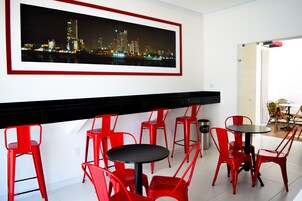Un área de comedor moderna con sillas rojas y mesas negras, un cuadro enmarcado con una vista urbana en la pared, y una pincelada de un área de comedor con sillas amarillas al fondo.