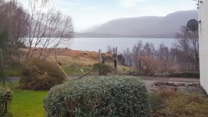 Blick auf einen See mit Bergen im Hintergrund, eine Wiese mit einem Busch und ein Gebäude mit Satellitenschüssel.