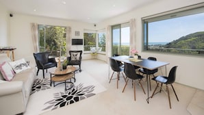 A spacious living room with a large window offering a scenic view, a dining area with modern furniture, and a cozy seating area with a sofa and armchairs. - Hilltop Getaway - Superb Sea & Mountain Views with Spa pool! (Nelson)