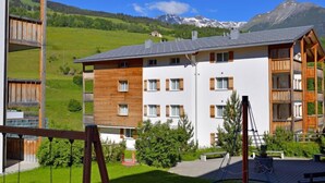 A multi-story building with a mix of wooden and white exterior walls, set against a backdrop of green hills and mountains. - Surses Alpin by Interhome (Savognin)