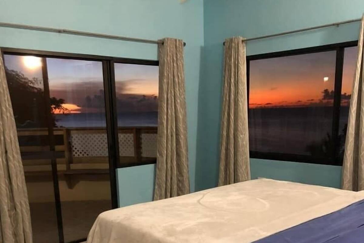 Une chambre à coucher avec un lit, une vue sur le coucher de soleil à travers de grandes fenêtres et un balcon avec une balustrade.