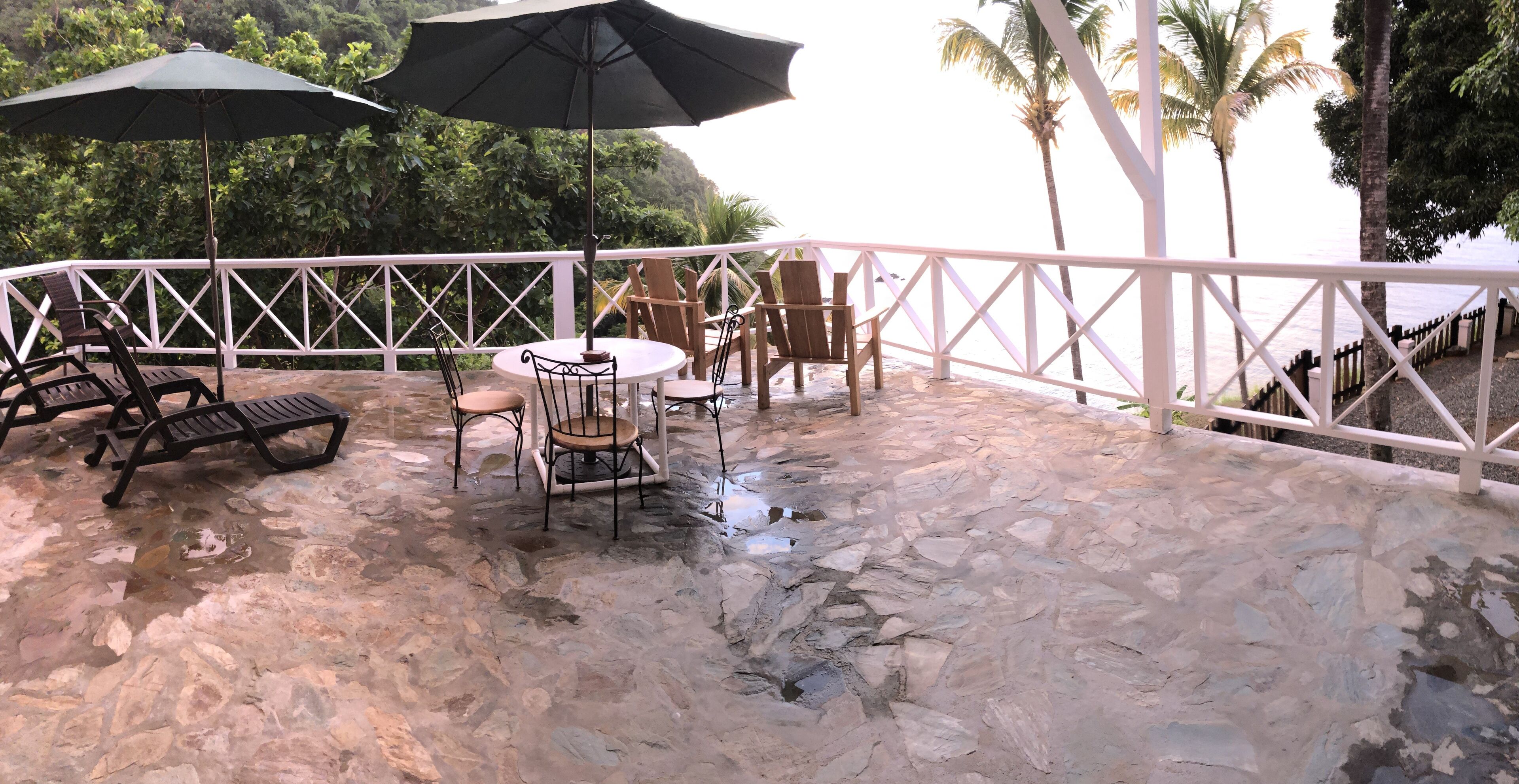 A stone patio with a table and chairs, two umbrellas, and a white railing.