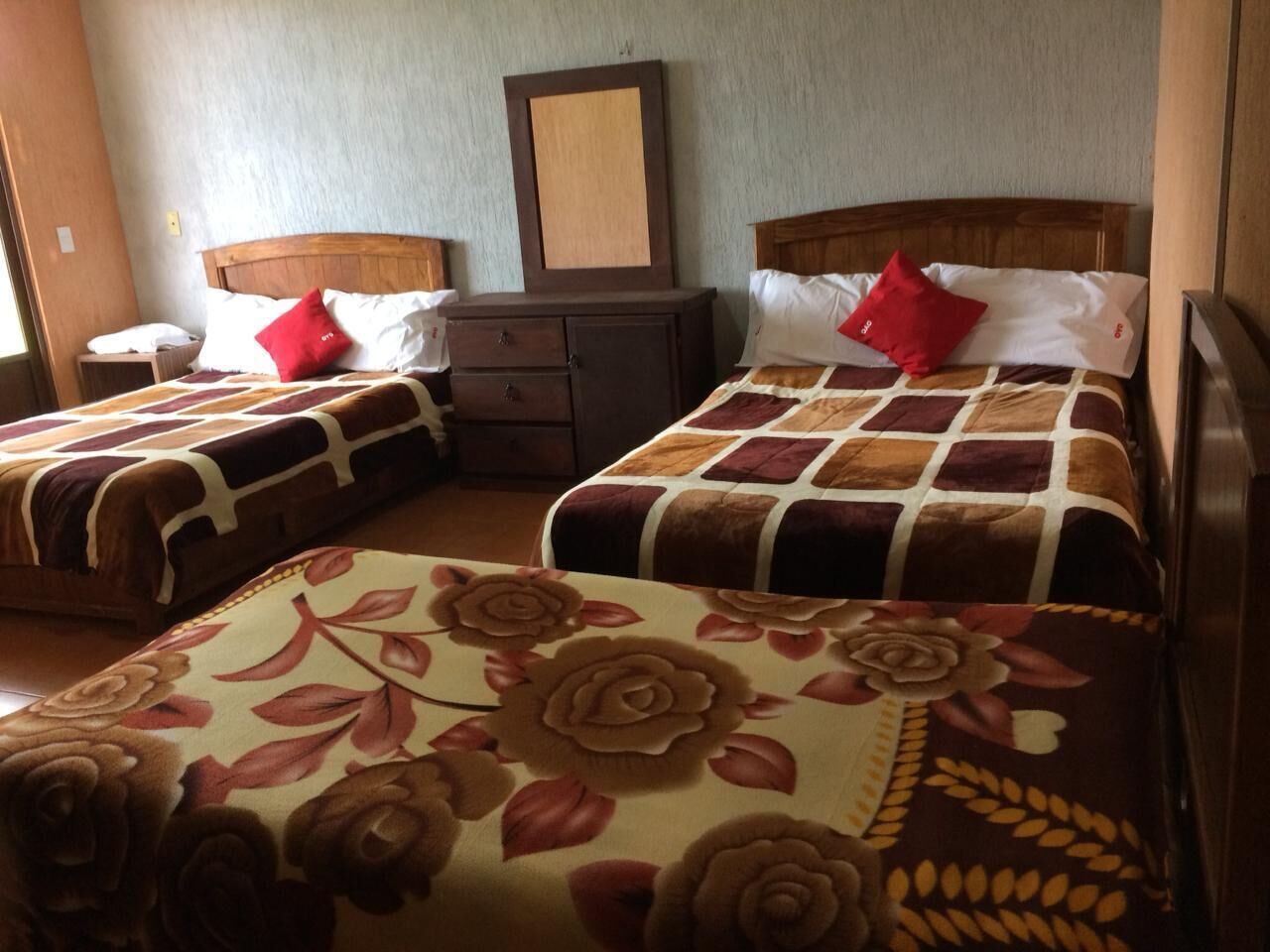 A hotel room with two single beds, each with a wooden headboard and floral-patterned bedding. There is a wooden nightstand with a lamp and a small table with a chair.