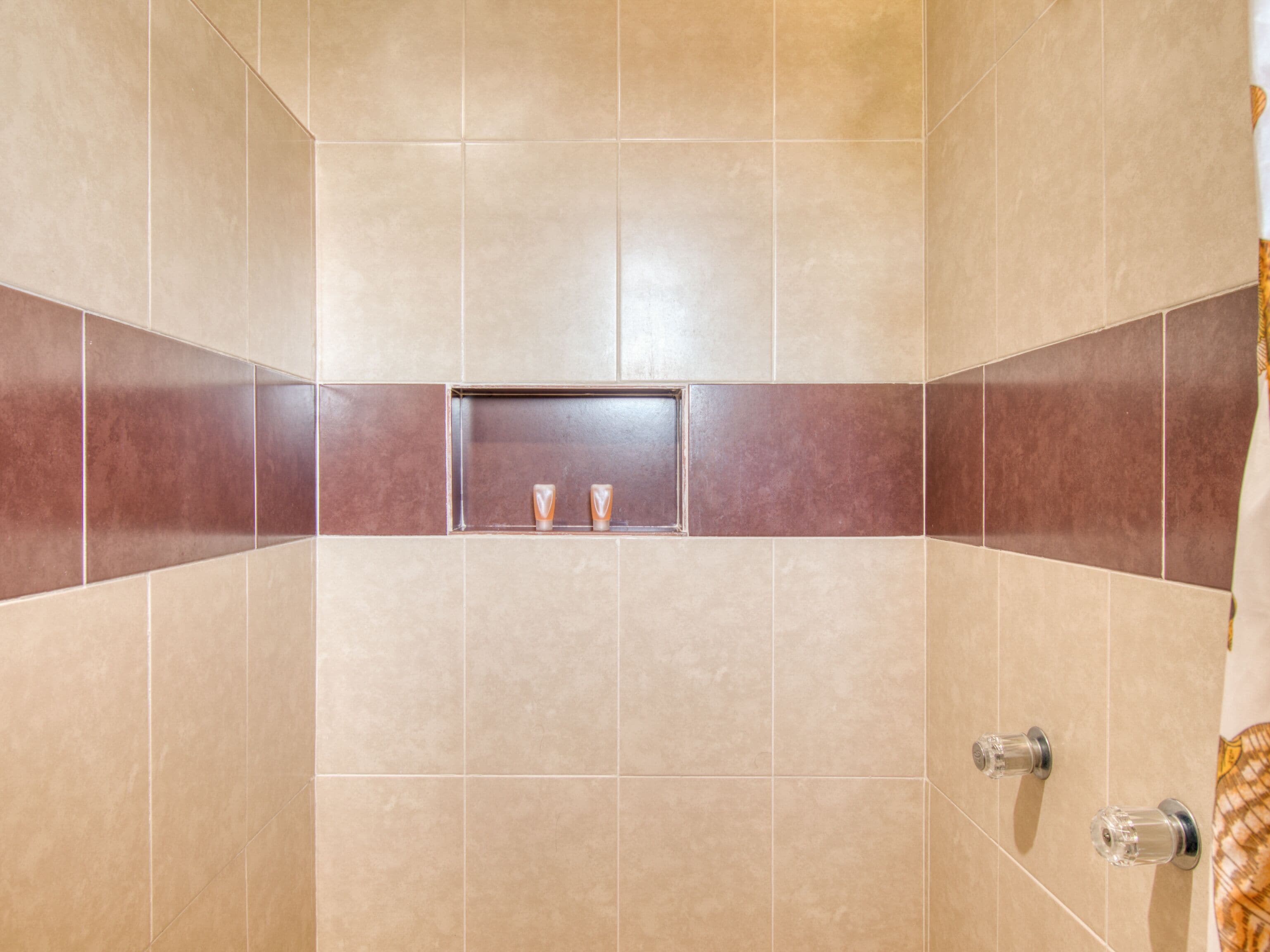 A bathroom with beige tiles, a central shelf with two bottles, and a shower curtain with a gold pattern.