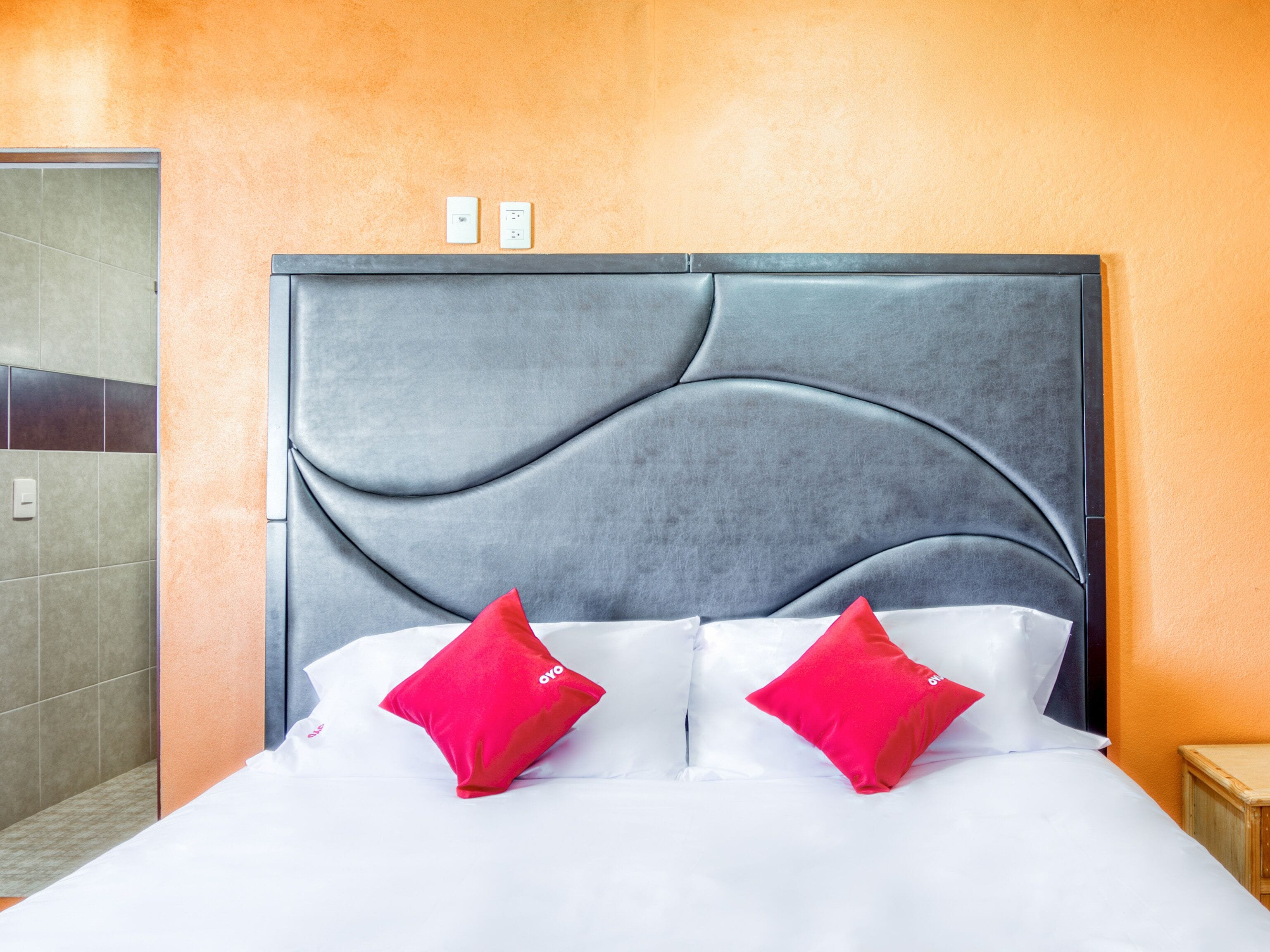 A hotel room with a grey upholstered headboard, white bedding, and two red pillows.
