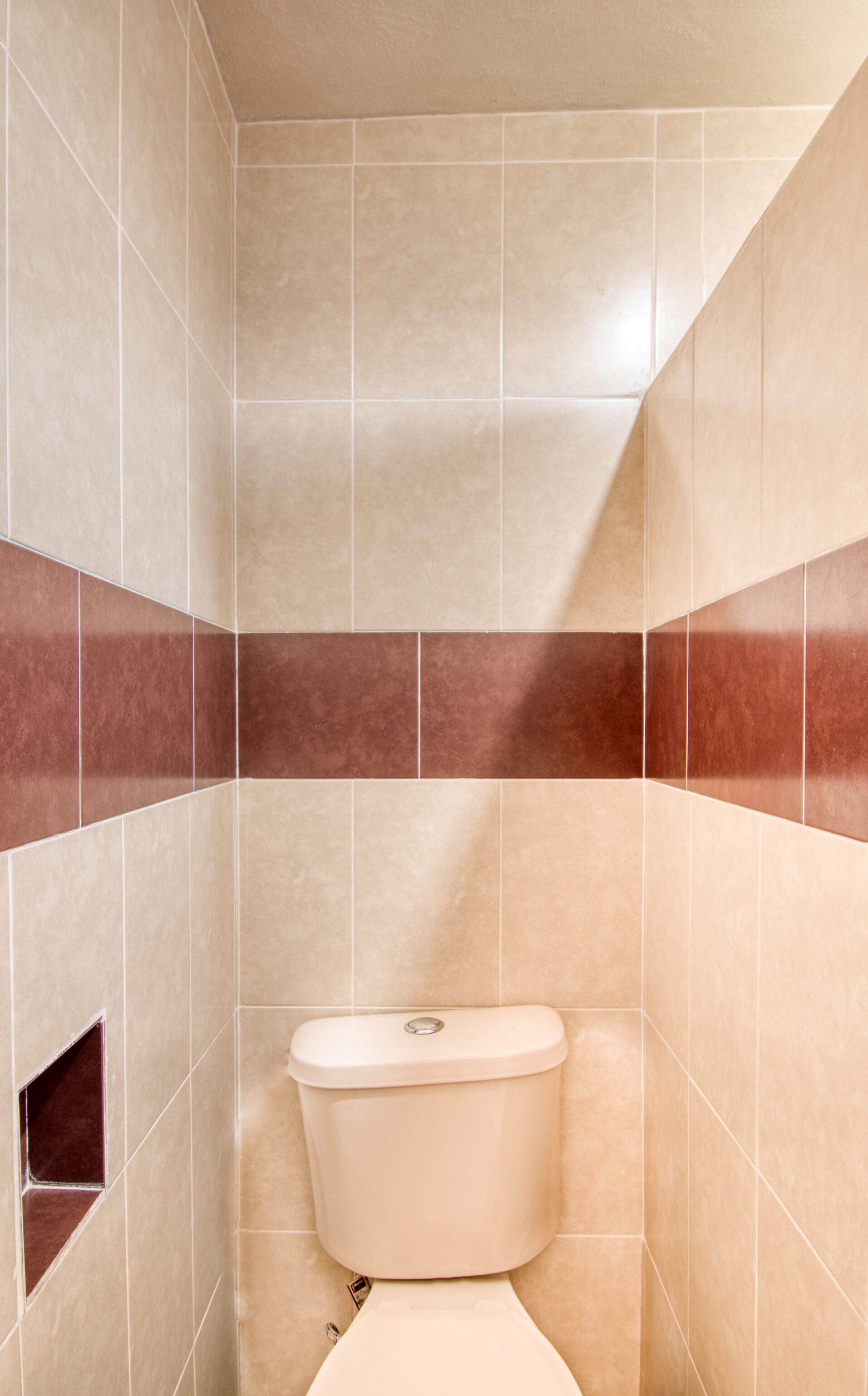 Ein kleines, schmales Badezimmer mit beige- und braunen Fliesen, einer weißen Toilette und einem kleinen Regal.