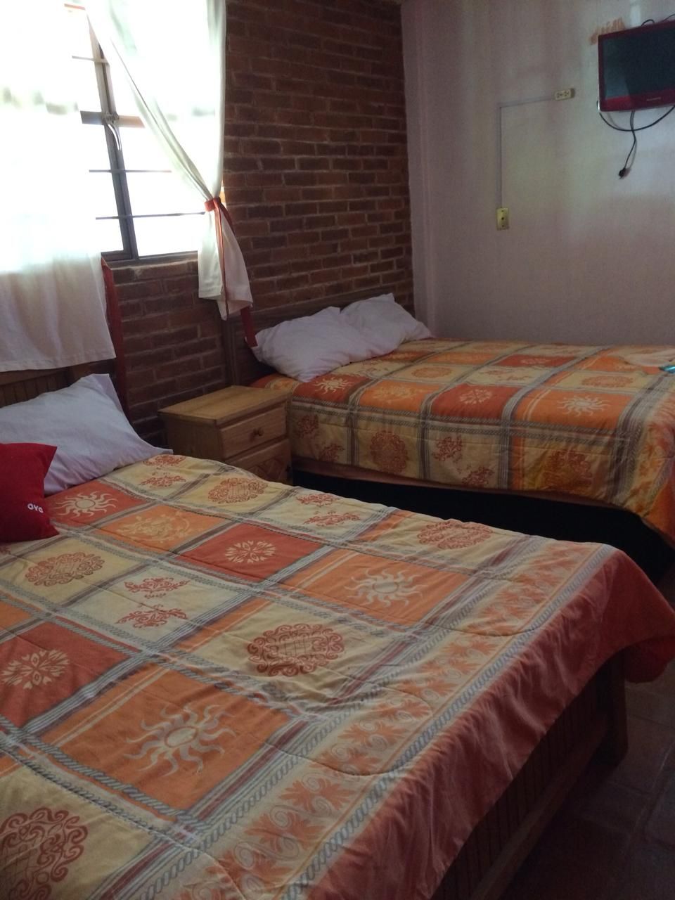 Una habitación con dos camas, cada una con una colcha estampada, una mesita de noche de madera, una pared de ladrillo y una ventana con cortinas.