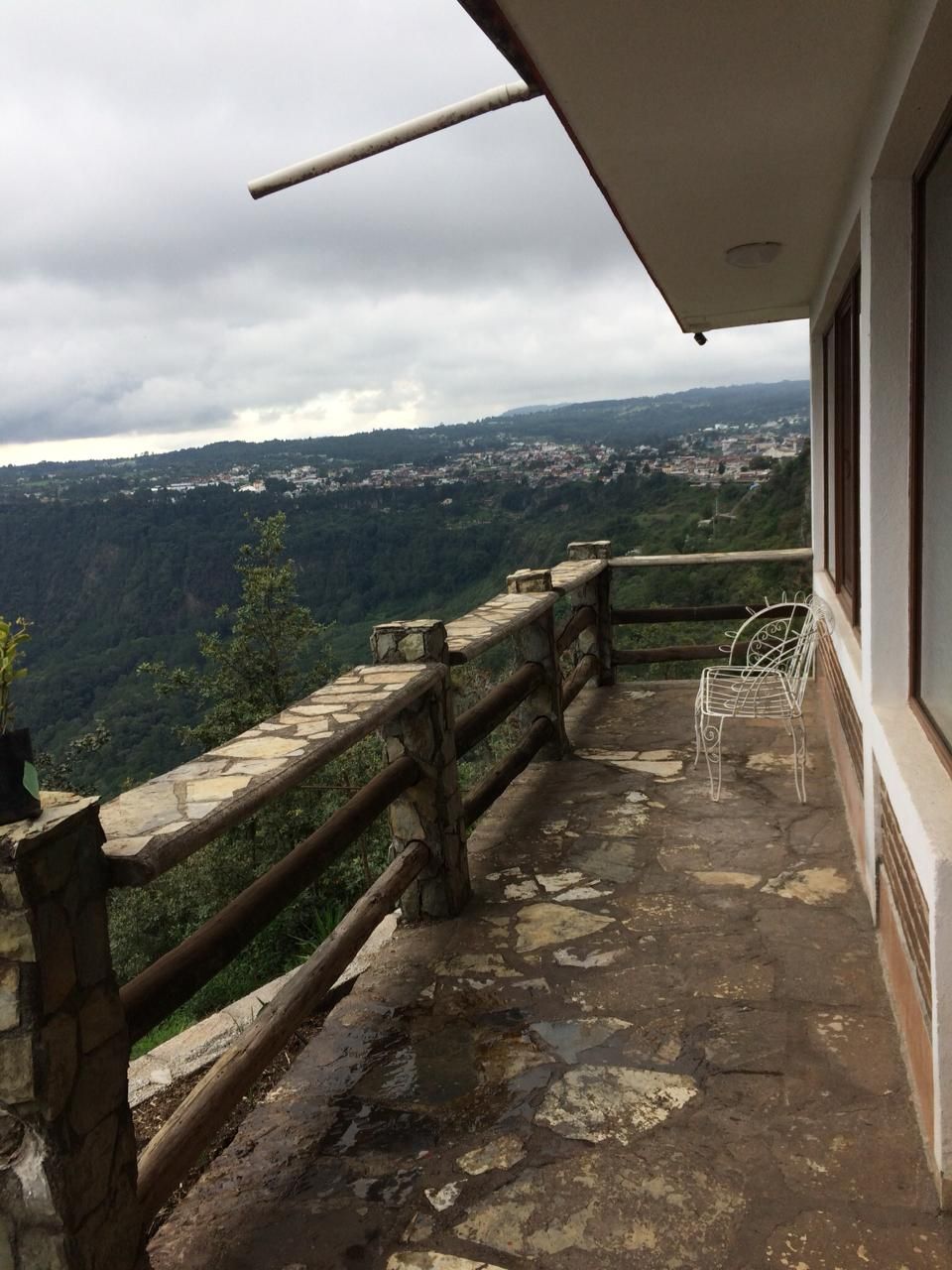 Un balcón con barandilla de piedra, una silla de metal y vistas a un pueblo y colinas boscosas.