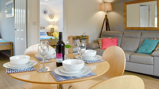 A dining area with a round table set for four, a bottle of wine, and a sofa in the background. - 2 bedroom accommodation in Chideock (Chideock)
