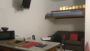 Un espacio habitable pequeño con kitchenette, un sofá, televisor montado en la pared y una cama nido.