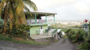 Una casa de dos plantas con balcón, rodeada de follaje y vistas a un pueblo.