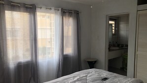 Una camera da letto con un letto, un comodino, un tavolo di appoggio, un bagno con lavandino e specchio, e una finestra con tende.