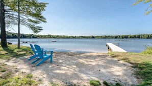 Blick auf einen See mit einem Steg, einem Sandstrand und blau gestreiften Adirondack-Stühlen, sowie eine bewaldete Uferlinie.