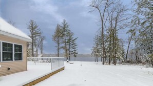 Ein Haus mit einem mit Schnee bedeckten Deck und Blick auf einen von Bäumen umgebenen See.