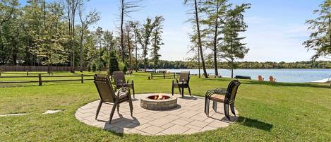 A circular outdoor seating area with a fire pit, surrounded by chairs, overlooking a lake.