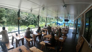 A covered outdoor seating area with wooden tables and benches, large windows offering views of greenery, and a ceiling fan.