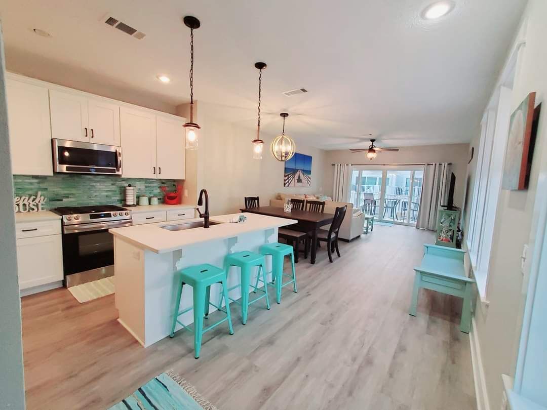 A modern kitchen with white cabinets, a central island with bar stools, a dining area with a table and chairs, and a view of the outdoors through a large window.